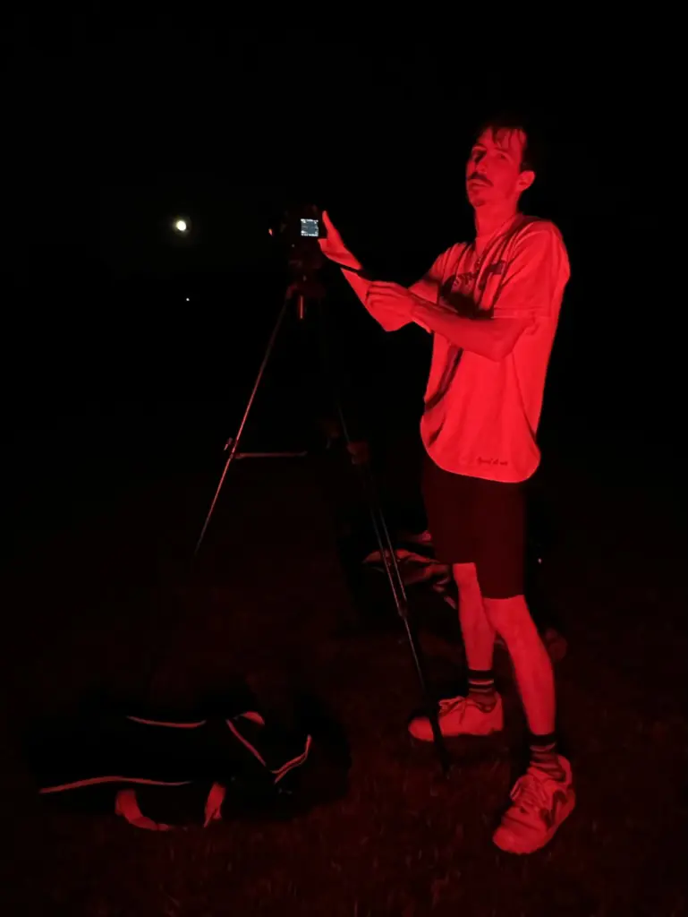Ben Armes, illuminated with red light, preparing a camera on a tripod in the dark.